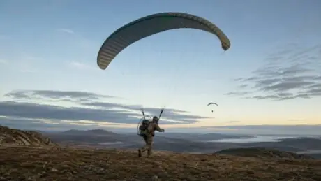 Original illustration of an anonymous service member launching a compact powered paraglider across a remote training landscape at dawn.